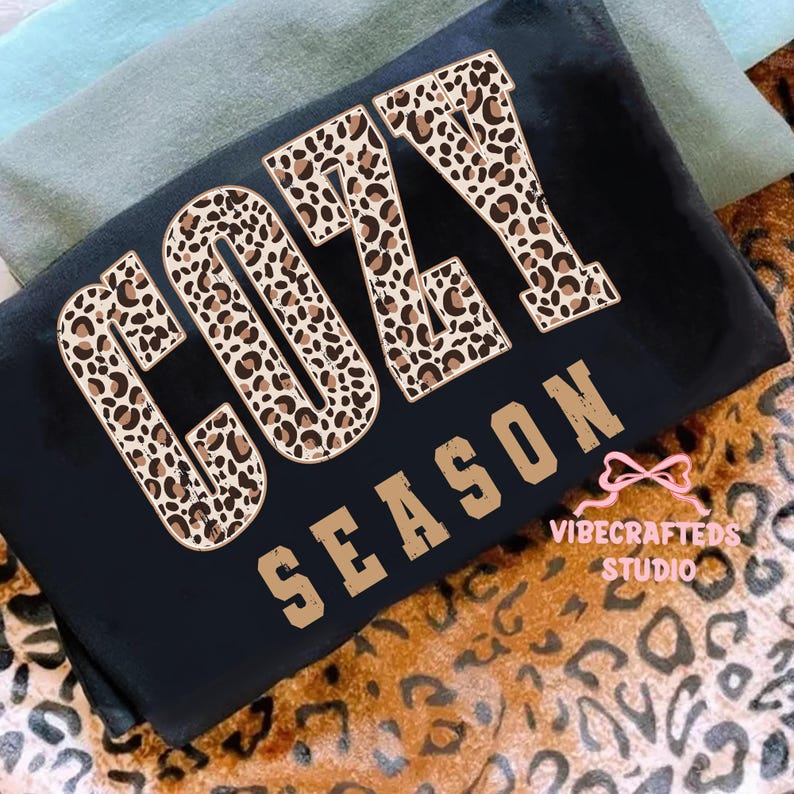 May include: Black t-shirt with the words "COZY SEASON" in a combination of leopard print and tan block letters. The shirt is folded and resting on a leopard print fabric.