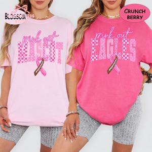 May include: Two short-sleeve t-shirts in light pink and dark pink. The light pink shirt features the text "pink out MASCOT" and the dark pink shirt features "pink out EAGLES" with a brown football-patterned ribbon graphic on each shirt.