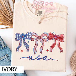 May include: Ivory t-shirt with three patriotic bows in red, white, and blue, and the word "USA" in blue script. The shirt has a crew neck and short sleeves. The shirt is made of a soft, comfortable material. The t-shirt is a size M.