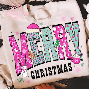May include: Cream-colored sweatshirt with a festive "MERRY CHRISTMAS" design. The text features pink and teal letters with holiday-themed patterns and illustrations, including a Santa hat, gingerbread man, and snowflakes.
