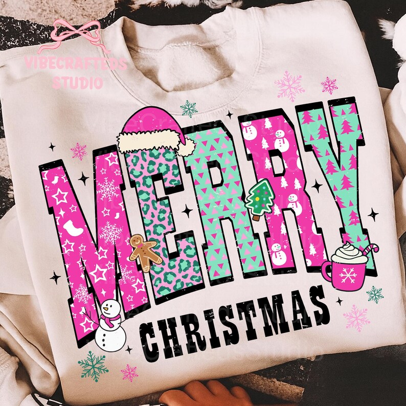 May include: Cream-colored sweatshirt with a festive "MERRY CHRISTMAS" design. The text features pink and teal letters with holiday-themed patterns and illustrations, including a Santa hat, gingerbread man, and snowflakes.