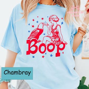May include: Pale blue t-shirt with a red graphic of an eagle, a woman, and the word "Boop." Red and blue stars are scattered around the design. The word "Chambray" is printed on a teal rectangle.