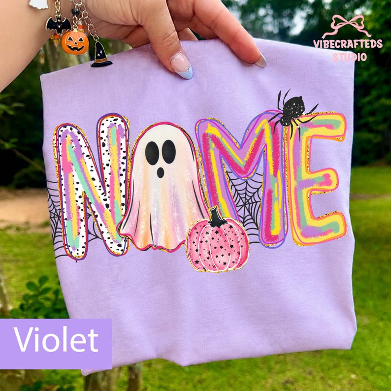 May include: A light purple t-shirt featuring a colorful graphic with the word NAME. The letter A is replaced by a ghost, and a pink pumpkin with black stars sits below. A black spider and web design are integrated into the lettering.