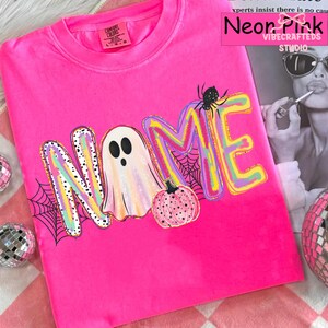 May include: A neon pink t-shirt featuring a graphic print with the word NAME, where the letter A is replaced by a white ghost. The design includes a pink polka-dot pumpkin, a black spider, and black spiderwebs. The shirt has a Comfort Colors label.