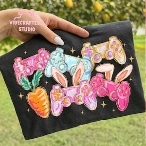Puede incluir: Camiseta gris oscuro con un gráfico colorido de mandos de videojuegos con orejas de conejo y una zanahoria. Los mandos son rosas, azules y naranjas con detalles dorados. El texto "VIBECRAFTED STUDIO" es visible.