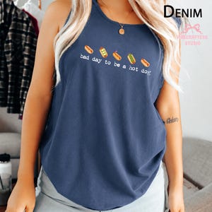 May include: A dark blue sleeveless tank top featuring a graphic of five small hot dogs arranged in a row above the white text that reads, bad day to be a hot dog. The shirt has a round neckline and a relaxed fit, showcasing a casual style.