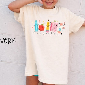 Puede incluir: Una camiseta de color marfil con un estampado gr&aacute;fico de material escolar antropom&oacute;rfico que incluye una cera azul, una manzana roja, un l&aacute;piz amarillo, un cuaderno rosa y una barra de pegamento, todos con caras sonrientes, junto al texto IVORY.