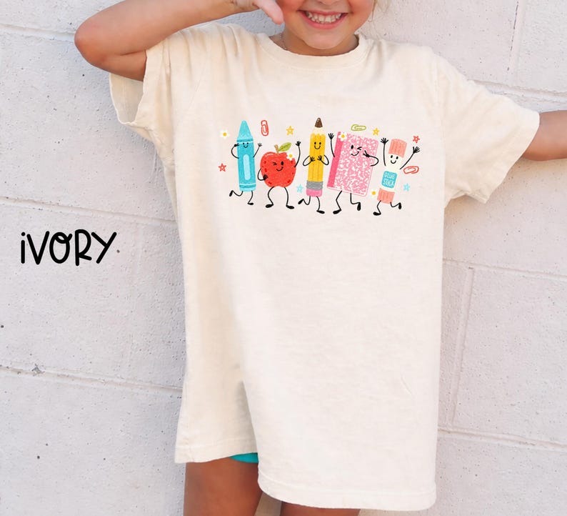 Puede incluir: Una camiseta de color marfil con un estampado gr&aacute;fico de material escolar antropom&oacute;rfico que incluye una cera azul, una manzana roja, un l&aacute;piz amarillo, un cuaderno rosa y una barra de pegamento, todos con caras sonrientes, junto al texto IVORY.