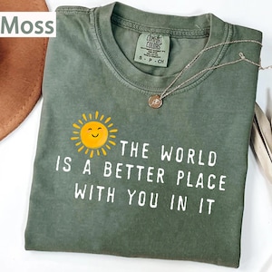 May include: A moss green short-sleeve t-shirt featuring a graphic of a yellow smiling sun and the white text "THE WORLD IS A BETTER PLACE WITH YOU IN IT". The shirt has a crew neckline and a Comfort Colors brand label inside the collar.