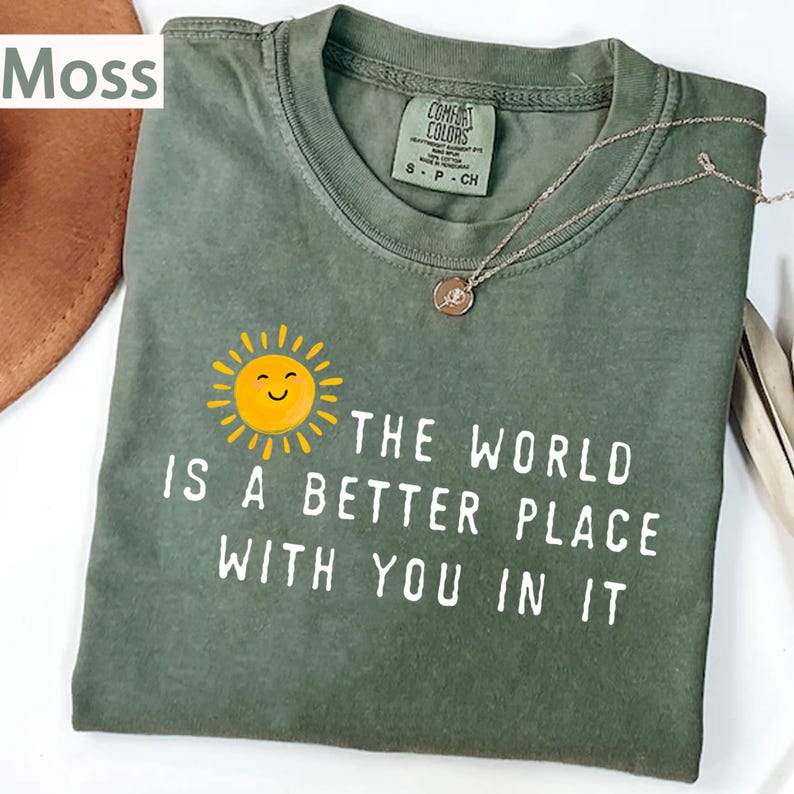 May include: A moss green short-sleeve t-shirt featuring a graphic of a yellow smiling sun and the white text "THE WORLD IS A BETTER PLACE WITH YOU IN IT". The shirt has a crew neckline and a Comfort Colors brand label inside the collar.