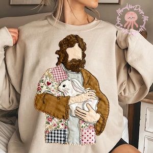May include: Beige sweatshirt with an embroidered design of a figure holding a white rabbit. The figure has brown hair and beard, with a patchwork design on the clothing. The patchwork includes floral, gingham, and polka dot patterns.