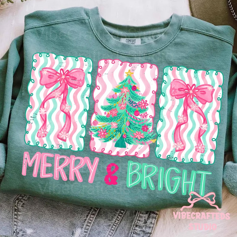 May include: A teal green sweatshirt with a festive design. The design includes three rectangular frames with pink bows and a Christmas tree. The words "MERRY & BRIGHT" are printed in pink and green. The sweatshirt is made of a soft material.