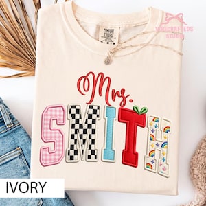 以下が含まれることがあります： アイボリー色のTシャツに「Mrs. SMITH」の刺繍。文字はピンクギンガム、白黒チェッカー、水色、緑の葉付き赤、虹をテーマにした様々なスタイルです。繊細なネックレスがシャツにかけられています。