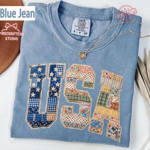 Puede incluir: Una camiseta azul claro con la palabra "USA" aplicada en la parte delantera. Las letras están hechas de tela de retazos en varios patrones y colores. La camiseta tiene cuello redondo y mangas cortas. La etiqueta de la marca dice "Comfort Colors".