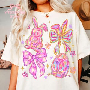 May include: Cream-colored t-shirt featuring colorful Easter-themed graphics. The design includes a pink bunny, bows, an egg, and flowers, all with glitter accents. The text "VIBECRAFTERS STUDIO" is visible in the upper left corner.