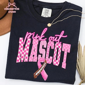 May include: A black t-shirt featuring the text "pink out" in neon pink script above the word "MASCOT" in large block letters. Each letter has a different pink pattern, including checkers, dots, stripes, and leopard print, with a football ribbon.