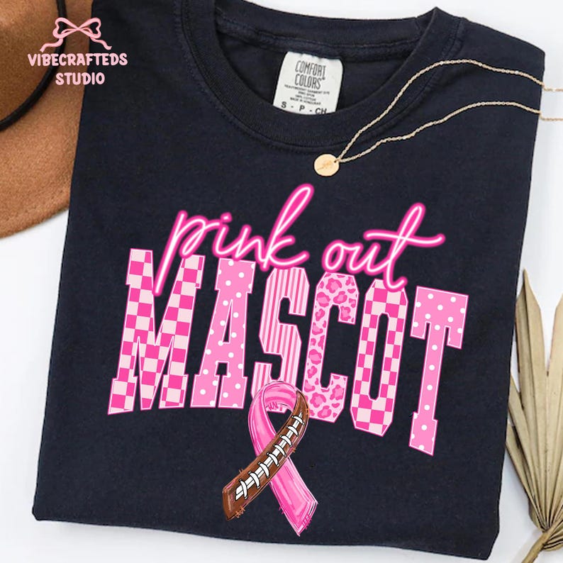 May include: A black t-shirt featuring the text "pink out" in neon pink script above the word "MASCOT" in large block letters. Each letter has a different pink pattern, including checkers, dots, stripes, and leopard print, with a football ribbon.