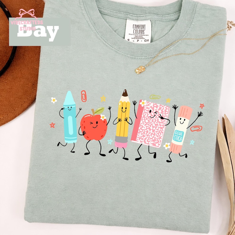 Puede incluir: Una camiseta verde salvia con un estampado gr&aacute;fico de cinco &uacute;tiles escolares antropom&oacute;rficos, incluyendo un cray&oacute;n azul, una manzana roja, un l&aacute;piz amarillo, un cuaderno rosa y un pegamento en barra, todos con caras sonrientes.