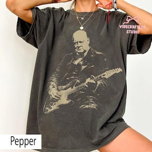 Peut inclure: T-shirt gris foncé avec un motif sépia d'un homme jouant de la guitare électrique. Le texte "Pepper" est en bas et "VIBECRAFTEDS STUDIO" en haut à droite. Le t-shirt a des manches courtes et une coupe ample.