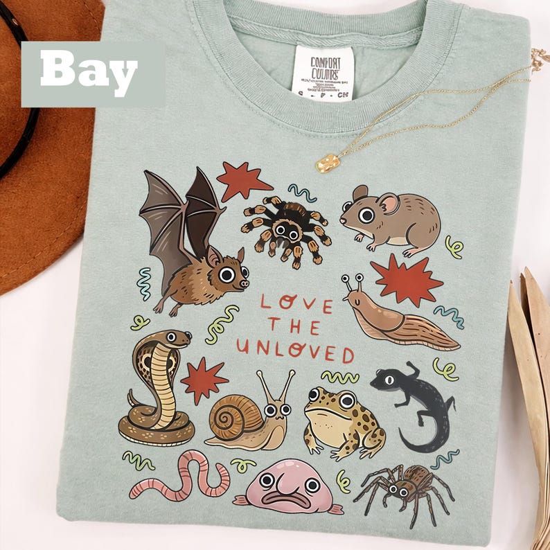 May include: A pale green Comfort Colors t-shirt featuring a collection of illustrated creatures such as a bat, spider, and snake. The words "LOVE THE UNLOVED" are printed on the front. A gold necklace is draped across the shirt.