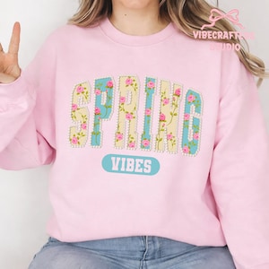 May include: A light pink sweatshirt with the word "SPRING" in a floral design. The letters are outlined with pearls and filled with a light blue and yellow pattern, adorned with pink roses. Below the word "SPRING" is the word "VIBES" in a light blue oval.