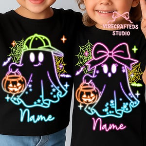 May include: Black t-shirts featuring neon ghost designs holding jack-o'-lanterns. One ghost wears a baseball cap, the other a bow. The word "Name" is written in cursive below each ghost. The shirts have spiderwebs and stars.