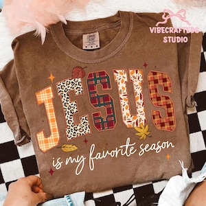 May include: Brown Comfort Colors t-shirt with the word "JESUS" in a fall-themed design. The letters feature plaid, leopard print, and leaf patterns. The phrase "is my favorite season" is written below.