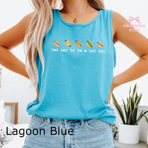 May include: A bright lagoon blue sleeveless tank top featuring a graphic of five small hot dogs in a row above the white text that reads, bad day to be a hot dog. The shirt has a round neckline and is worn with light blue denim jeans.