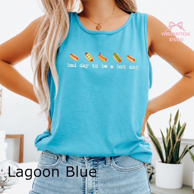 May include: A bright lagoon blue sleeveless tank top featuring a graphic of five small hot dogs in a row above the white text that reads, bad day to be a hot dog. The shirt has a round neckline and is worn with light blue denim jeans.