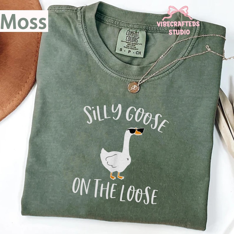 May include: Moss green t-shirt with the words "Silly Goose On The Loose" and a cartoon goose wearing sunglasses. The shirt has a crew neck and short sleeves. A silver necklace with a small pendant is on the shirt.