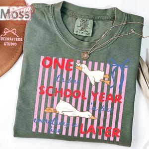 May include: Sage green Comfort Colors t-shirt with the text "One to for School Year I'm fine everything is later" in red and blue. The design includes two cartoon geese and pink vertical stripes. A silver necklace with a pendant is on the shirt.