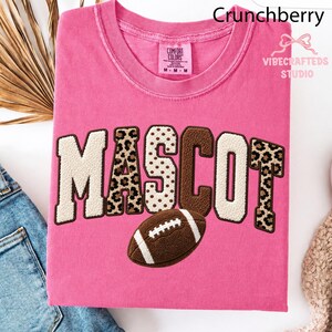 May include: A pink t-shirt featuring the word MASCOT in large, textured letters with a mix of cream, brown, and leopard print patterns. A brown football graphic is positioned below the text. The shirt has a Comfort Colors brand label.