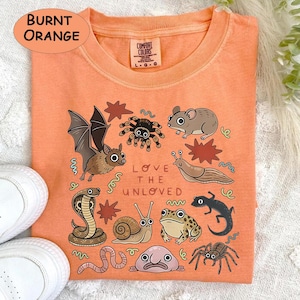 May include: Burnt orange Comfort Colors sweatshirt with a vibrant illustration of creatures like a bat, spider, mouse, and snake. The text "LOVE THE UNLOVED" is printed below the illustrations.