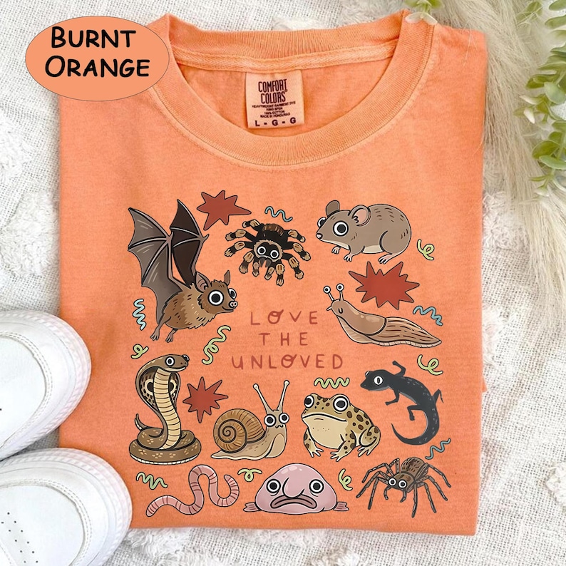 May include: Burnt orange Comfort Colors sweatshirt with a vibrant illustration of creatures like a bat, spider, mouse, and snake. The text "LOVE THE UNLOVED" is printed below the illustrations.