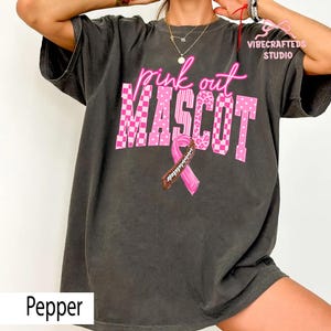 May include: A charcoal gray t-shirt featuring the pink text "pink out" above the word "MASCOT" in a large, blocky font with checkered, striped, and leopard patterns. A pink awareness ribbon with a brown football lace design is centered below.