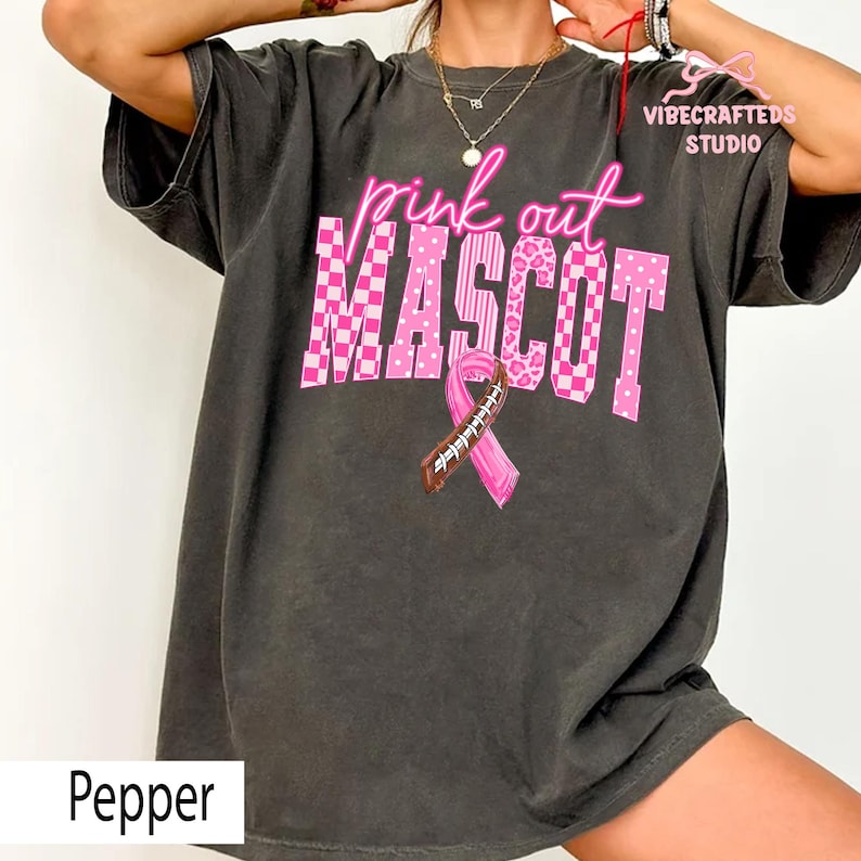 May include: A charcoal gray t-shirt featuring the pink text "pink out" above the word "MASCOT" in a large, blocky font with checkered, striped, and leopard patterns. A pink awareness ribbon with a brown football lace design is centered below.