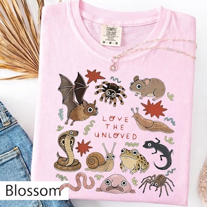 May include: Pale pink t-shirt featuring a collection of illustrated creatures such as a bat, spider, and snake, with the text "LOVE THE UNLOVED". A delicate necklace with a pendant is also visible. The t-shirt is labelled "Blossom".