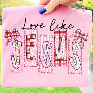 May include: Pink t-shirt with the words "Love like Jesus" in black script. The word "Jesus" is spelled out in decorative letters with hearts, gingham, and stripes. Red and white gingham bows adorn the top of the letters.