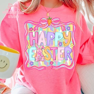 May include: Pink sweatshirt with a colorful "Happy Easter" graphic. The design includes a bunny, flowers, and a bow with a carrot. The text is in pastel colors, perfect for Easter celebrations.