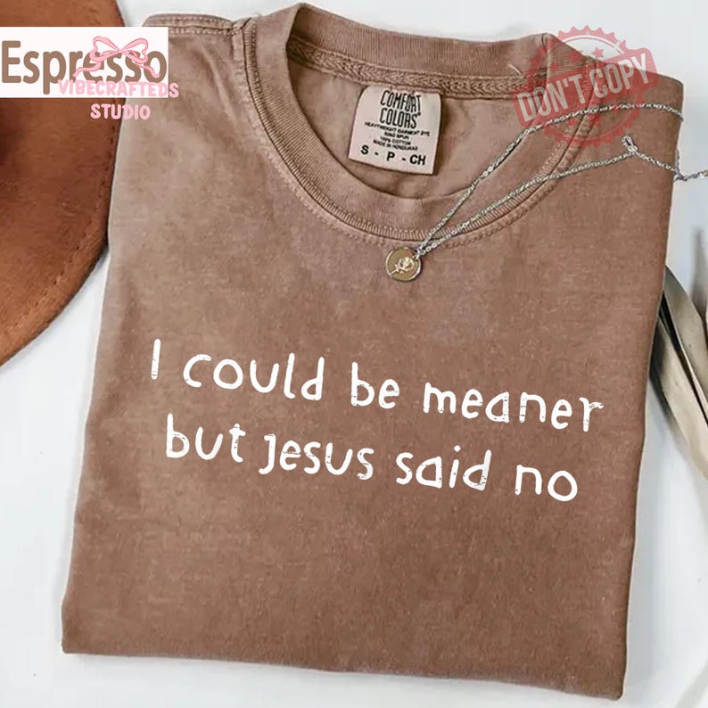 May include: A brown Comfort Colors t-shirt with the text "I could be meaner but Jesus said no" in white. A silver necklace with a pendant rests on the shirt. The shirt has a crew neck.