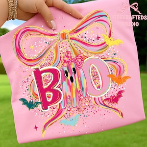 May include: Pink t-shirt with a colorful bow and the word "BOO" in a glittery font. A ghost with a bow and bats are also featured in the design. The shirt has a festive Halloween theme.