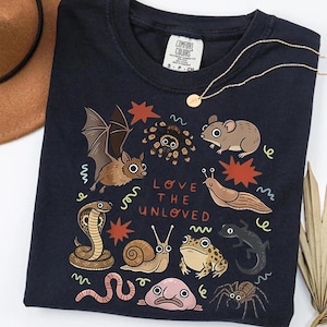 May include: Navy blue t-shirt with a colourful illustration of a bat, spider, and other creatures. The text "LOVE THE UNLOVED" is displayed below the illustrations. A gold necklace is draped across the shirt.