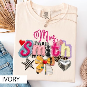 Pode incluir: Uma t-shirt de manga curta na cor marfim com o texto Mrs. Smith em letra cursiva rosa e letras coloridas e texturizadas. O design inclui uma estrela xadrez preta e branca, um laço com padrão de lápis amarelo e um coração prateado.