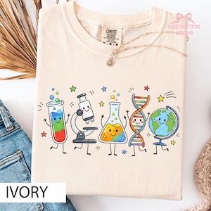 May include: An ivory t-shirt featuring a colorful graphic print of cartoon science icons including a test tube, microscope, flask, DNA strand, and globe, all with smiling faces, surrounded by small stars on a light-colored fabric background.