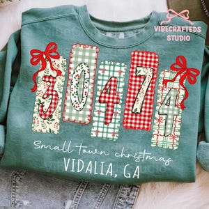 Pode incluir: Uma camisola verde-azulado com o texto "30474" num design festivo. Os números estão em padrões vermelhos e brancos, com laços vermelhos. As palavras "Small town christmas VIDALIA, GA" estão impressas abaixo.