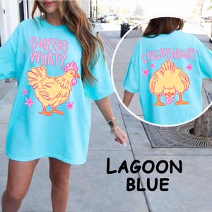 May include: Lagoon blue t-shirt with a playful chicken graphic. The front features the text "GUESS WHAT?" above a yellow chicken with pink accents and stars. The back displays a chicken's rear with the text "CHICKENBUTT".