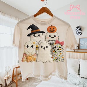 Op de afbeelding: Crèmekleurig T-shirt met een groep cartoongeesten met Halloween-thema's. Eén geest draagt een heksenhoed, een ander heeft een pompoen op zijn hoofd en anderen hebben strikken en patronen. Het shirt hangt aan een houten hanger.