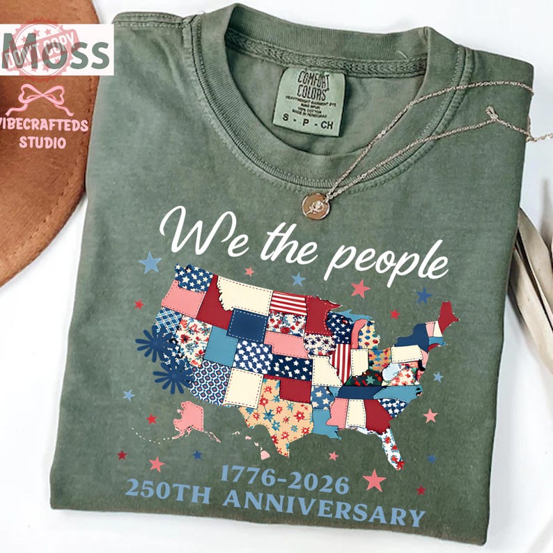 May include: An olive green t-shirt featuring the text "We the people" and a patchwork map of the United States. The shirt celebrates the 250th anniversary, with the years 1776-2026 printed below the map. A silver necklace with a pendant is on the shirt.