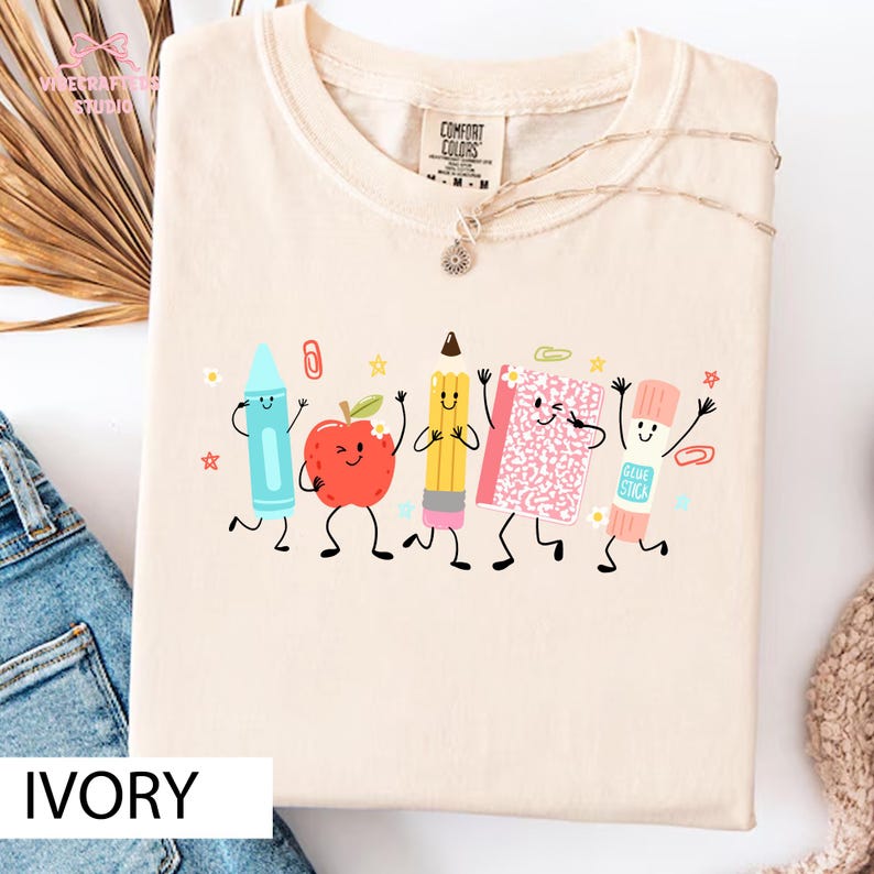Puede incluir: Una camiseta de color marfil con un estampado gr&aacute;fico de cinco &uacute;tiles escolares antropom&oacute;rficos, incluyendo un cray&oacute;n azul claro, una manzana roja, un l&aacute;piz amarillo, un cuaderno rosa y un pegamento en barra, todos con caras sonrientes.