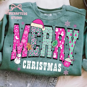 May include: A teal green sweatshirt with the words "MERRY CHRISTMAS" in large, colorful letters. The word "MERRY" is in pink with white stars, snowflakes, and a gingerbread man. The word "CHRISTMAS" is in white.